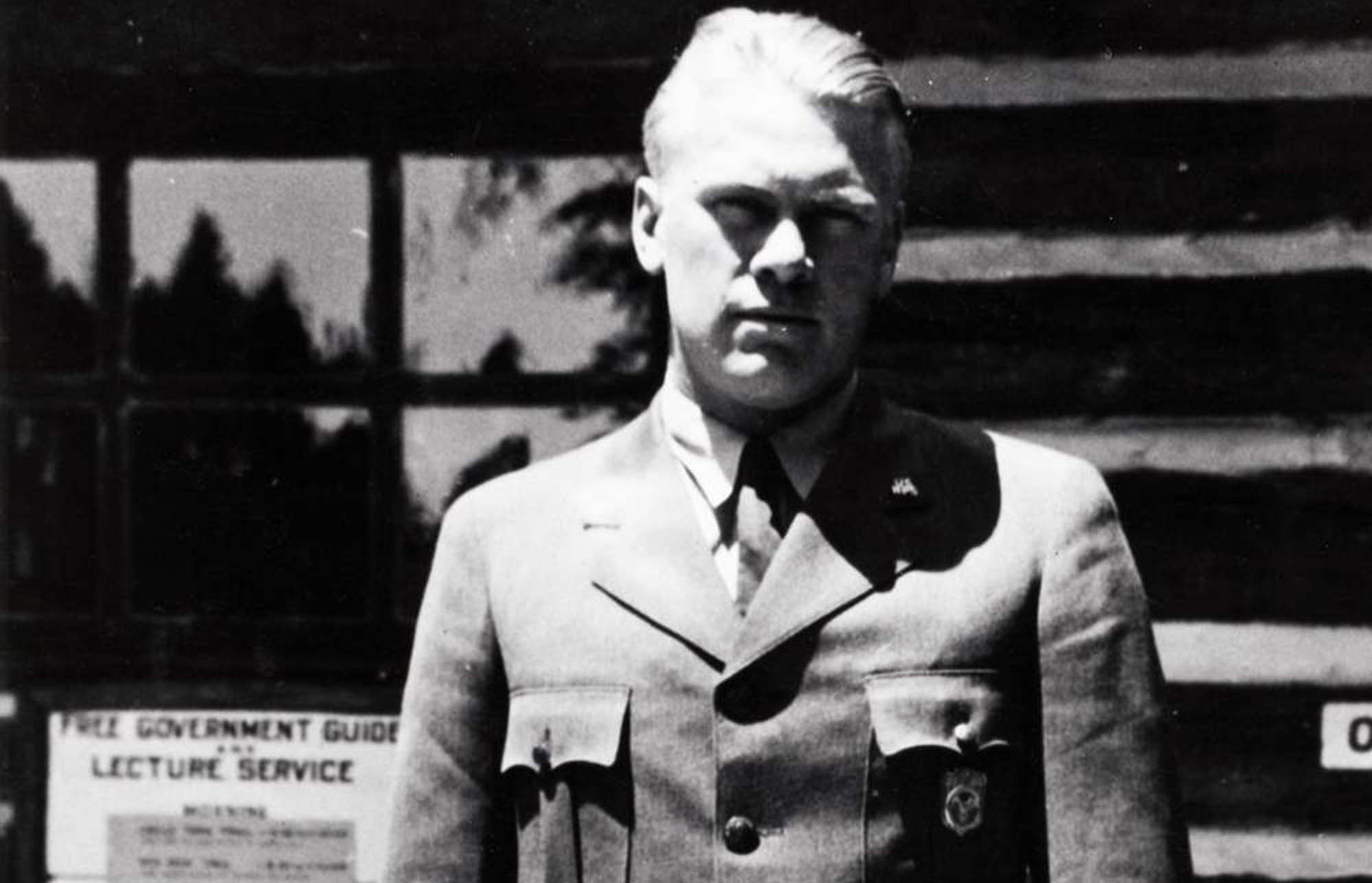 Gerald Ford poses in front of a log building while wearing the NPS uniform with shield-shaped badge and holding a broad brim hat.